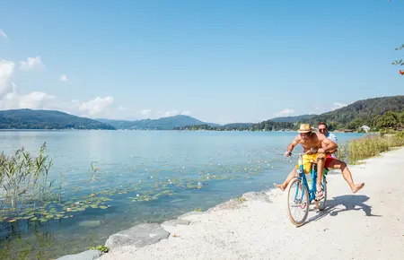 Zwei Personen sitzen auf einem Fahrrad und fahren am See entlang